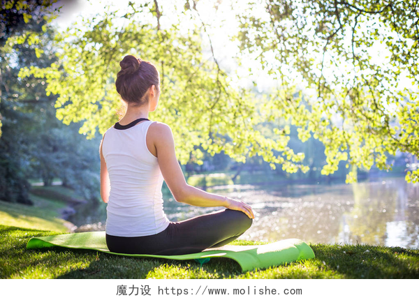 早晨练习瑜伽的女人背影年轻女子打坐, 而坐在瑜伽毯在上午公园附近的湖, 在街头锻炼的概念  